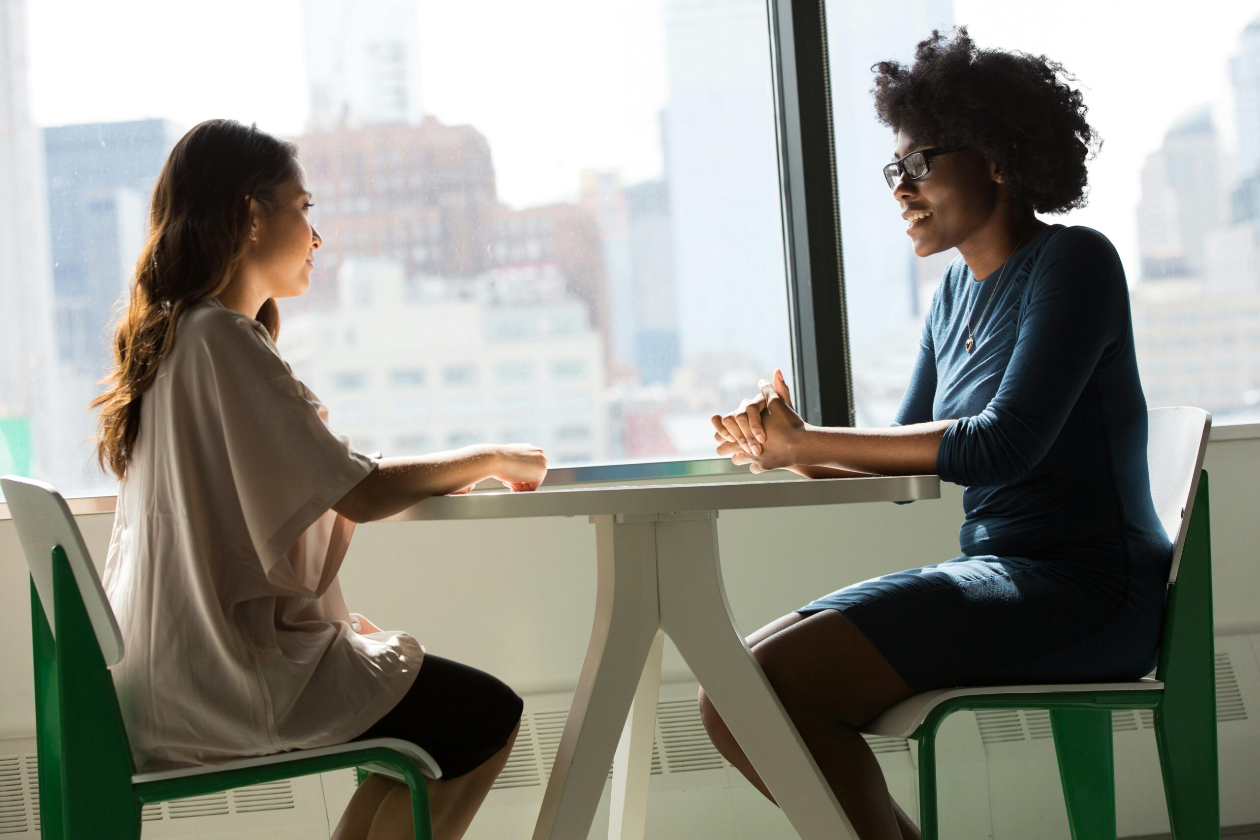 Two women sitting and talking at a table with a city view from the window.